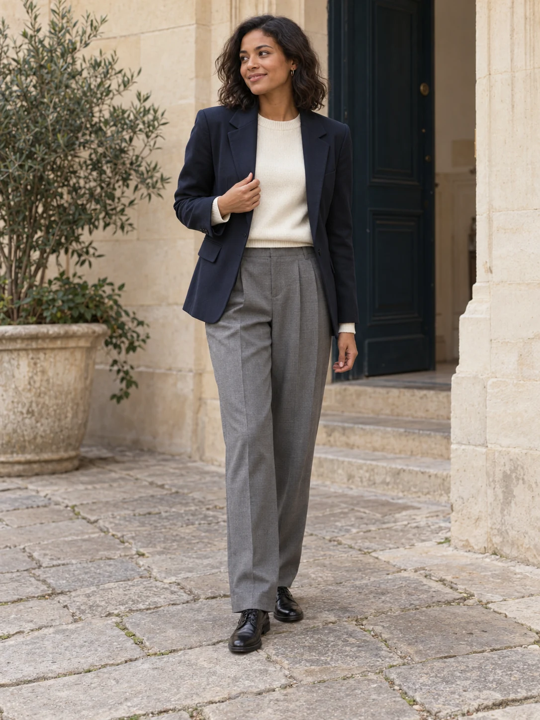 Une femme adulte porte un blazer marine ouvert sur une maille fine écrue à col rond, un pantalon gris à pinces et des derbies noires.